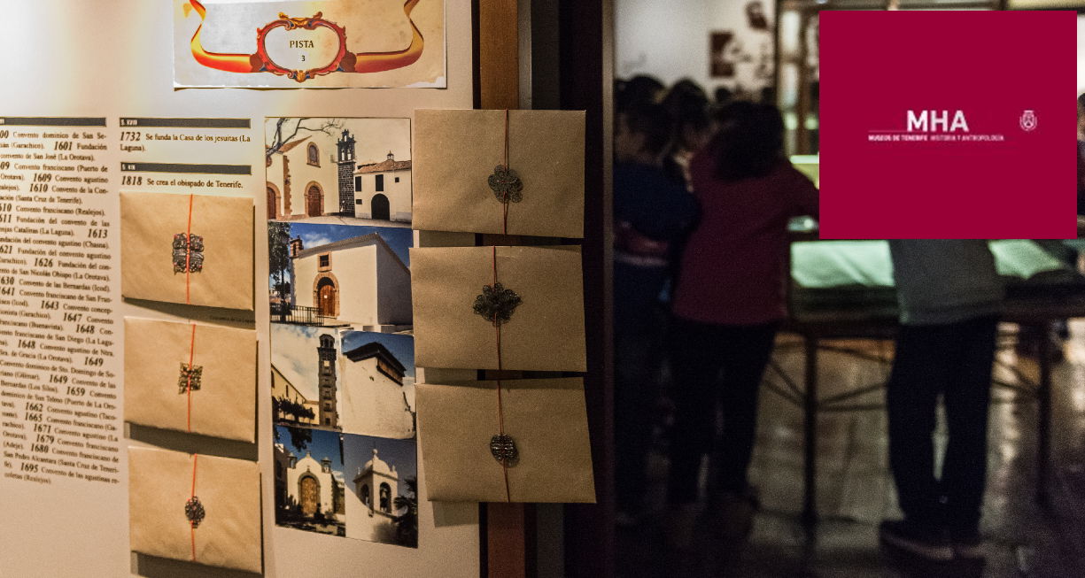 Instantánea tomada durante la realización del kit educativo. Muestra una serie de sobres colocados sobre uno de los paneles del museo y al fondo un grupo de niños. Con logo